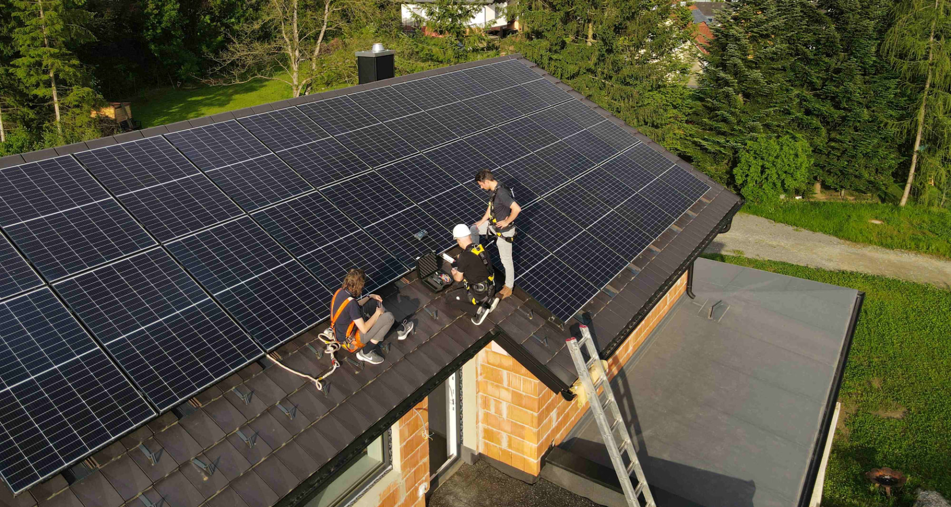 Luftaufnahme eines Satteldachs mit dunklen Photovoltaik-Modulen, auf dem drei Techniker mit Sicherungsgurten eine Anlagenprüfung durchführen